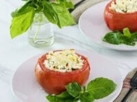 Square image of stuffed tomato on a white plate next to fresh basil leaves. Another plate, a sprig of basil in a glass of water, and a white towel with red stripes in the background.