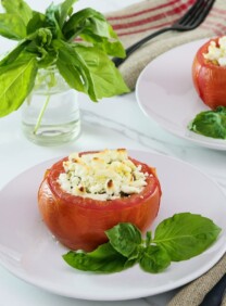 Square image of stuffed tomato on a white plate next to fresh basil leaves. Another plate, a sprig of basil in a glass of water, and a white towel with red stripes in the background.
