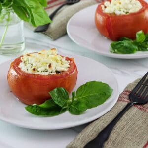 Horizontal image - stuffed tomato on a white plate next to fresh basil leaves, a napkin and fork lay to the right side. Another plate, a sprig of basil in a glass of water, and a white towel with red stripes in the background.