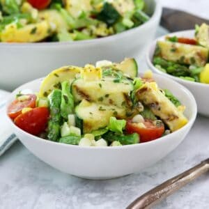 Horizontal image - Two white bowls filled with grilled vegetable salad, a mix of grilled zucchini, squash, asparagus, corn, tomatoes, lettuce and basil with lemony basil dressing. A fork sits off to the right side, a large bowl of the same salad in the background.