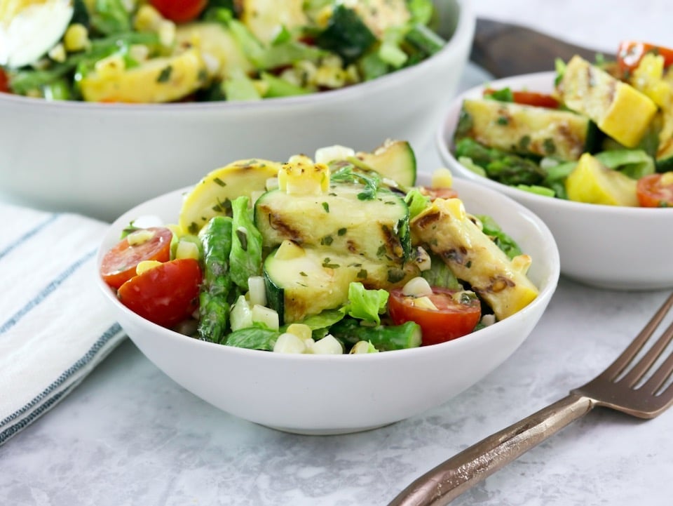 Horizontal image - Two white bowls filled with grilled vegetable salad, a mix of grilled zucchini, squash, asparagus, corn, tomatoes, lettuce and basil with lemony basil dressing. A fork sits off to the right side, a large bowl of the same salad in the background.