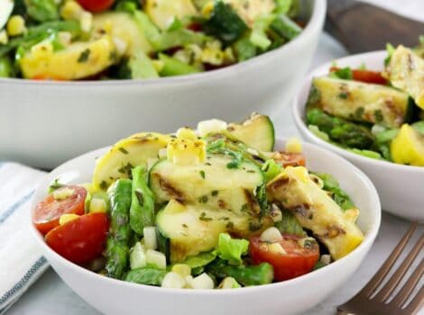 Square image - Two white bowls filled with grilled vegetable salad, a mix of grilled zucchini, squash, asparagus, corn, tomatoes, lettuce and basil with lemony basil dressing. A fork sits off to the right side, a large bowl of the same salad in the background.