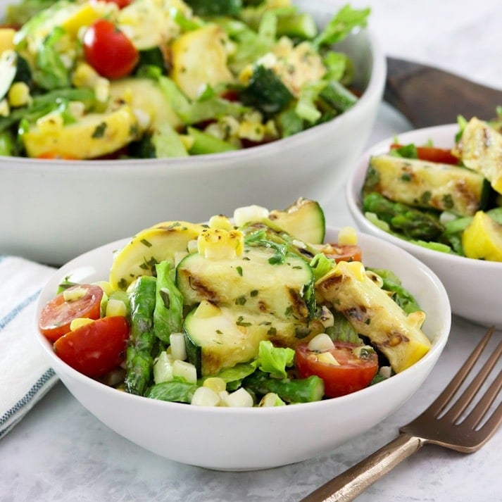 Square image - Two white bowls filled with grilled vegetable salad, a mix of grilled zucchini, squash, asparagus, corn, tomatoes, lettuce and basil with lemony basil dressing. A fork sits off to the right side, a large bowl of the same salad in the background.