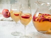 Horizontal shot of two wine glasses containing peach sangria and sliced peaches next to a glass pitcher of peach sangria and sliced peaches.