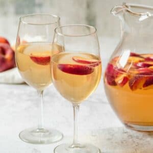 Horizontal shot of two wine glasses containing peach sangria and sliced peaches next to a glass pitcher of peach sangria and sliced peaches.