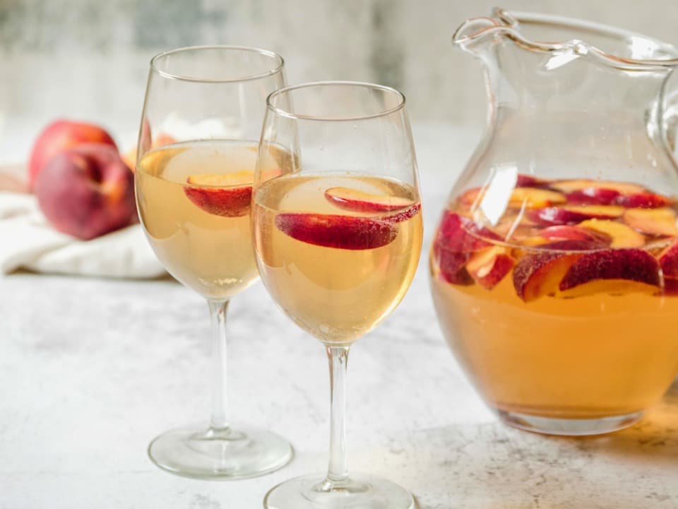 Horizontal shot of two wine glasses containing peach sangria and sliced peaches next to a glass pitcher of peach sangria and sliced peaches.