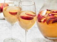 Close up shot of two wine glasses containing peach sangria and sliced peaches next to a glass pitcher of peach sangria and sliced peaches.