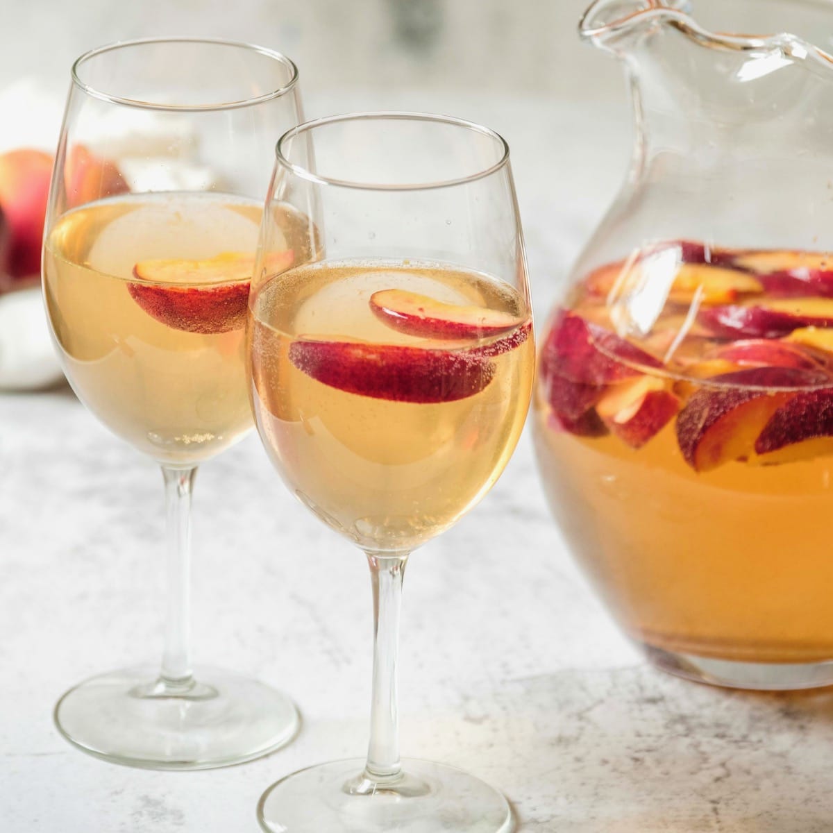Close up shot of two wine glasses containing peach sangria and sliced peaches next to a glass pitcher of peach sangria and sliced peaches.