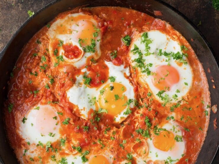 Horizontal close up image - cast iron pan filled with red shakshuka, topped with six cooked eggs and a sprinkle of fresh green herbs.