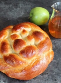 Round braided Apple Honey Challah on concrete background with two green apples and glass carafe of honey.