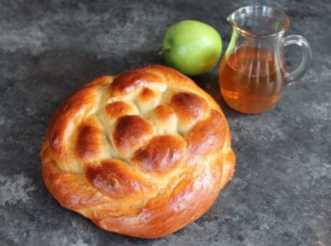 Round braided Apple Honey Challah on concrete background with two green apples and glass carafe of honey.