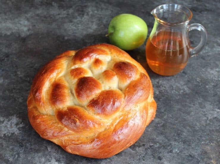 Round braided Apple Honey Challah on concrete background with two green apples and glass carafe of honey.