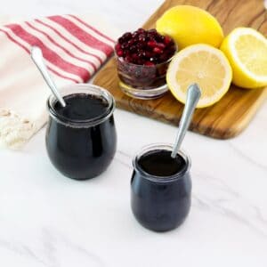 Horizontal shot of two glass jars filled with pomegranate molasses next to a cutting board holding sliced lemons and pomegranate arils.