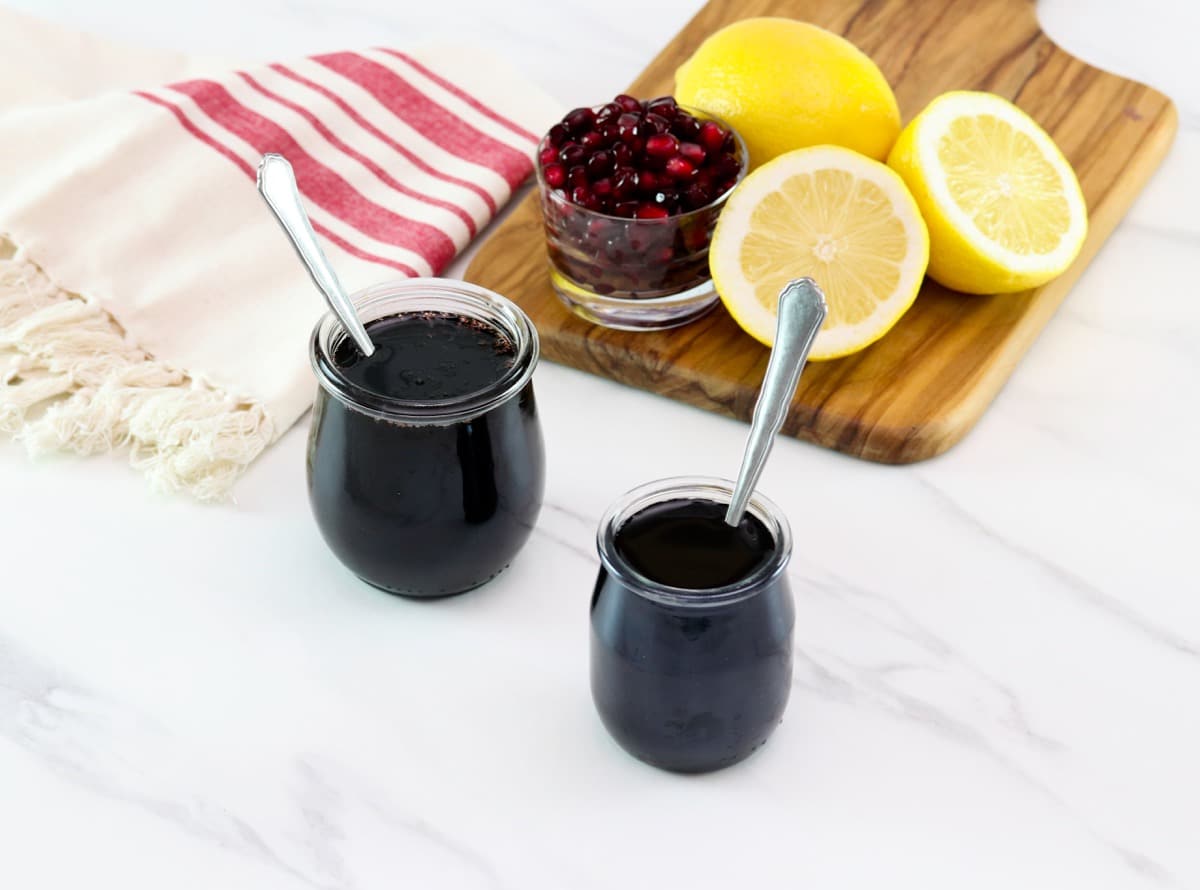 Horizontal shot of two glass jars filled with pomegranate molasses next to a cutting board holding sliced lemons and pomegranate arils.