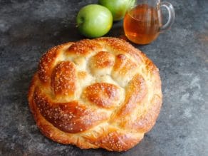 Round braided Apple Honey Challah with sparkling sugar coating on concrete background with two green apples and glass carafe of honey.