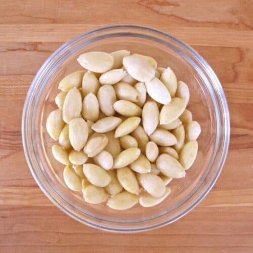 Blanched skinned almonds in glass bowl on wooden cutting board.
