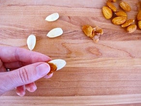 Fingers squeezing skin from almond, three skinned almonds and a small pile of almonds with skin on in the background.