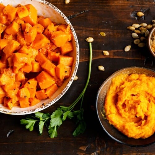 Overhead shot - a large bowl of tender roasted butternut squash cubes, a medium bowl filled with butternut squash puree, and a small bowl filled with toasted butternut squash seeds, on a dark wooden background. Fresh parsley and toasted butternut squash seeds are scattered beside the bowl.