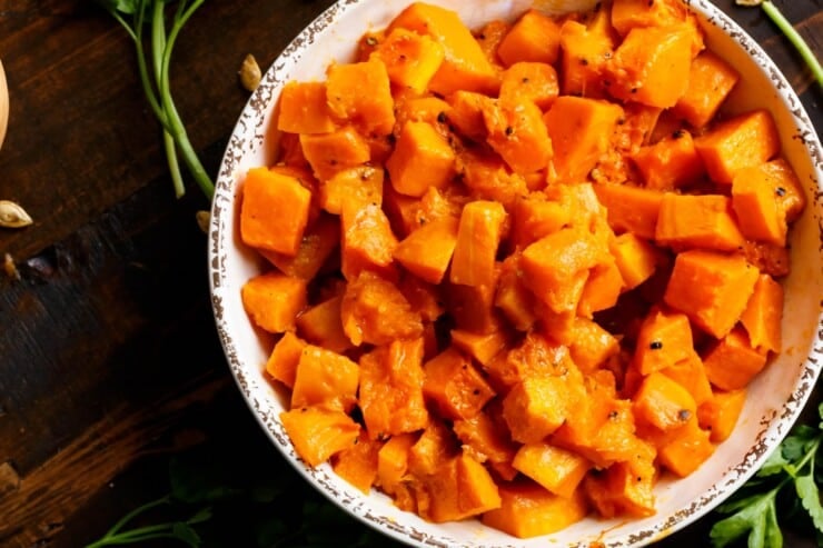 Overhead shot of a bowl filled with tender roasted butternut squash cubes on a dark wooden background, with fresh parsley and toasted butternut squash seeds scattered beside the bowl.