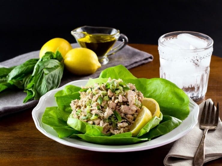 Healthy tuna salad on bed of green butter lettuce with lemon slices, on white plate with fork and cloth napkin, glass of ice water, lemons basil and olive oil in background.
