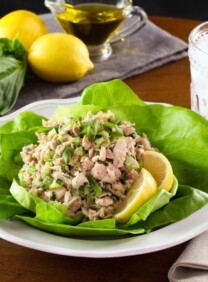 Healthy tuna salad on bed of green butter lettuce with lemon slices, on white plate with fork and cloth napkin, glass of ice water, lemons basil and olive oil in background.