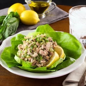 Healthy tuna salad on bed of green butter lettuce with lemon slices, on white plate with fork and cloth napkin, glass of ice water, lemons basil and olive oil in background.