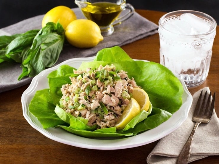 Healthy tuna salad on bed of green butter lettuce with lemon slices, on white plate with fork and cloth napkin, glass of ice water, lemons basil and olive oil in background.