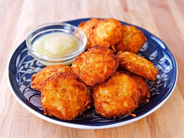 Pile of golden latkes on blue plate with applesauce on wooden background.