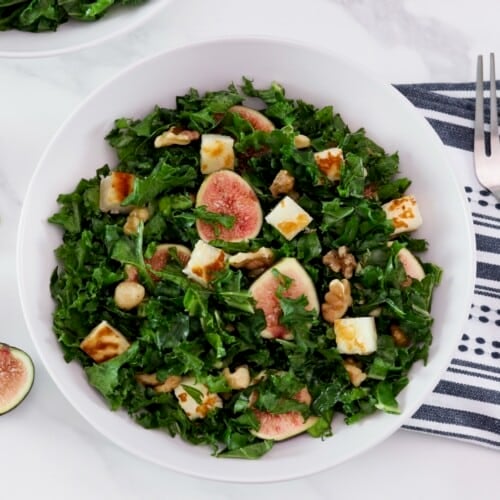 Overhead shot - large white bowl filled with fig salad with dark green curly kale, pan-seared halloumi cheese, and walnuts. Fork and linen napkin beside the bowl, fresh fig halves scattered across the marble countertop.