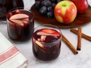 Tilted overhead shot of two glasses of red wine Thanksgiving sangria with fruit slices, three cinnamon sticks on the counter, a tray of fruit - apples and grapes, and a pitcher of sangria in the background. Linen towel on the side.