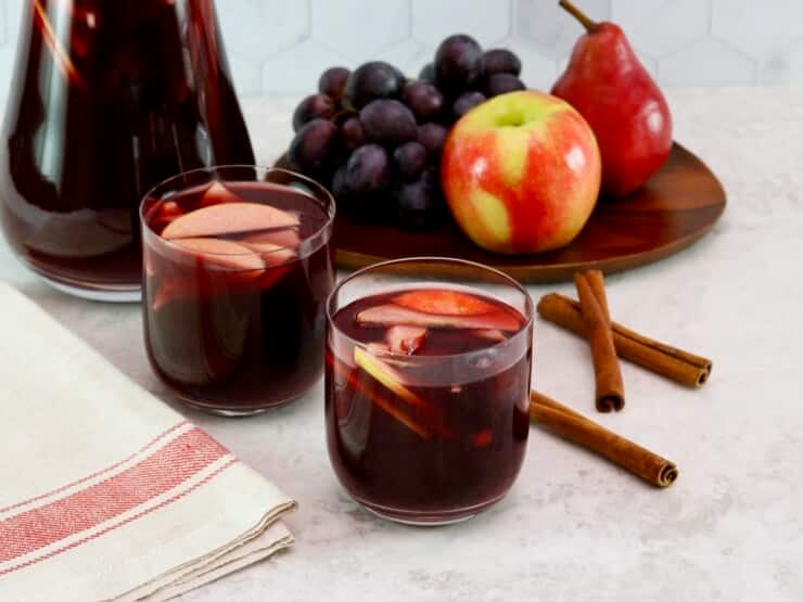 Close up shot - two unstemmed wine glasses filled with red wine Thanksgiving sangria, fresh fruit slices - apples and grapes floating in the cups. Linen towel beside the cups. On a white marble countertop with three cinnamon sticks, a platter of fruit - apple, grapes, pears - and a large pitcher of sangria in the background.