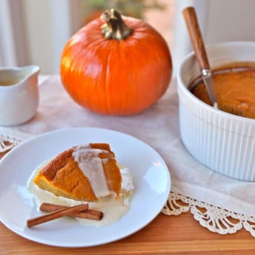 A serving of Pumpkin Pudding on a white small plate accompanied with two cinnamon sticks and a background a Pumpkin Pudding on a baking container and a whole pumpkin