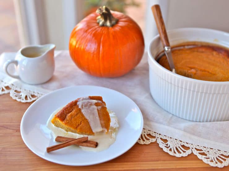 A serving of Pumpkin Pudding on a white small plate accompanied with two cinnamon sticks and a background a Pumpkin Pudding on a baking container and a whole pumpkin