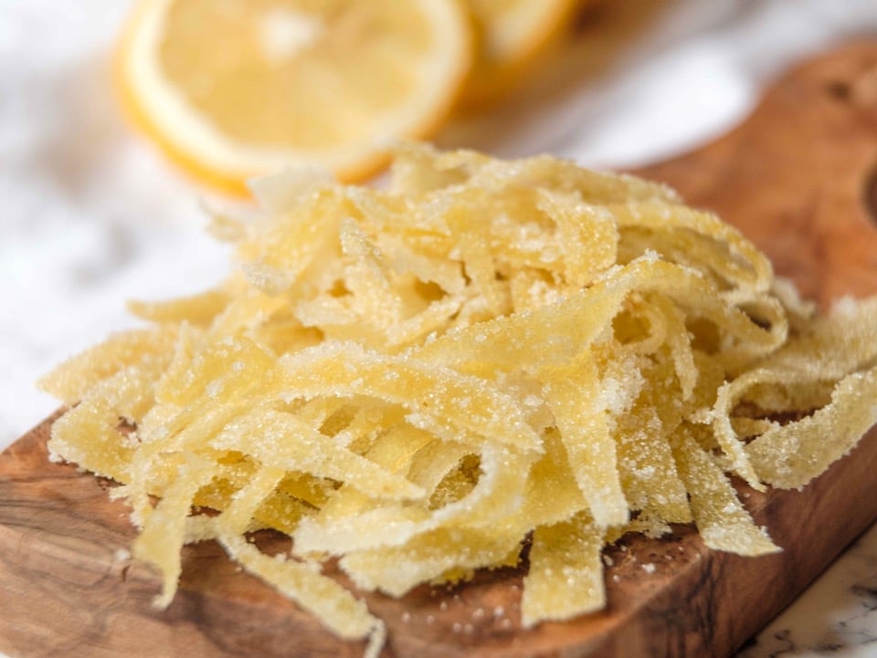 Close up of thin, dainty candied lemon peels coated in sugar - very thin ribbons of peel on a wooden cutting board, white linen napkin and lemon half in background, on a white marble surface.