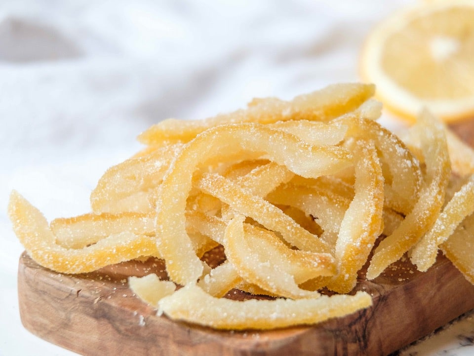 Close up of thick candied lemon peels coated in sugar - thick slices of peel on a wood cutting board, white linen napkin and lemon half in background, on a white marble surface.