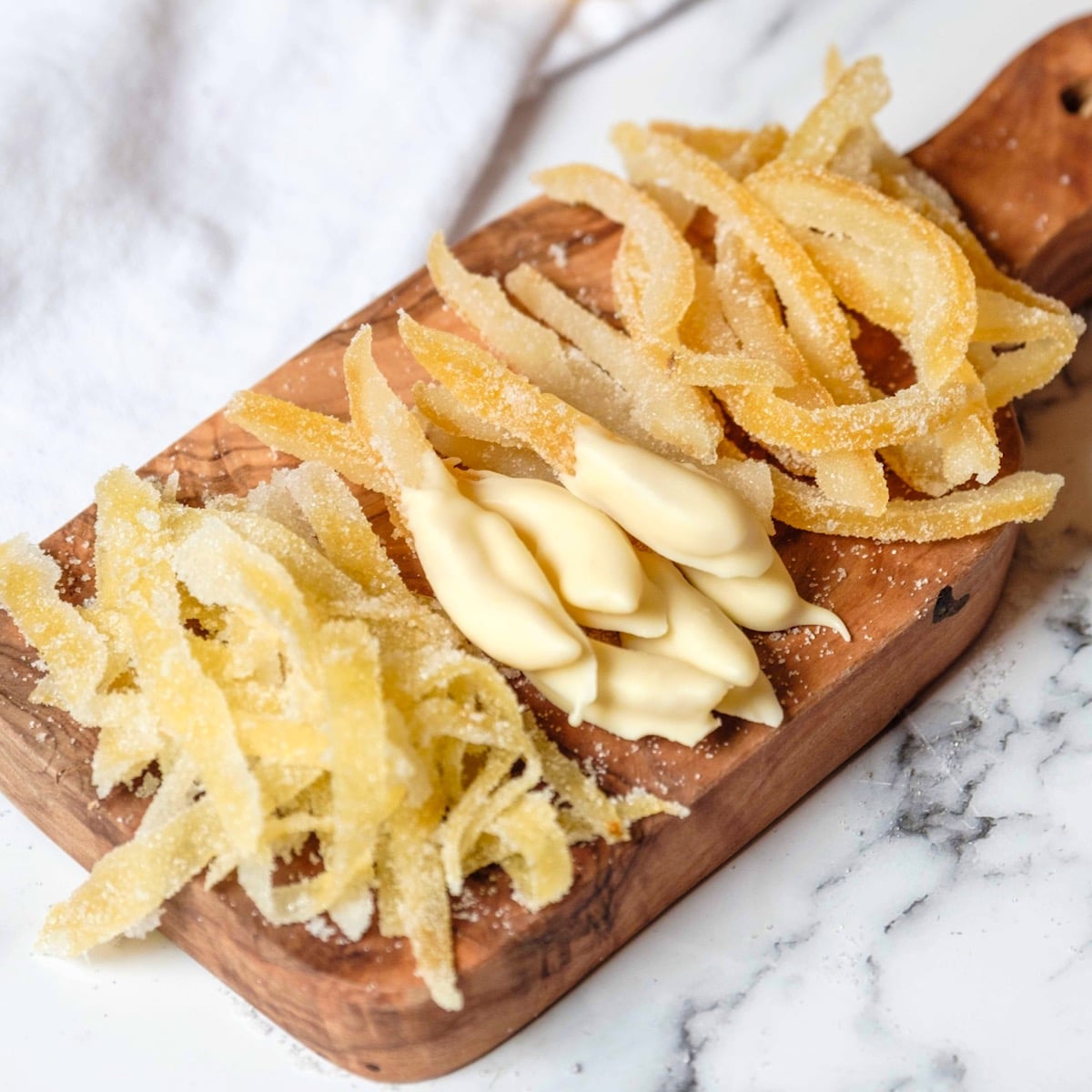 Featured square shot - wide shot of candied lemon peels on a small wood cutting board on marble counter. Large peels, small peels, peels dipped in white chocolate. White linen towel in background.