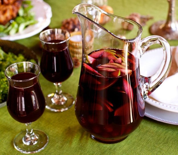 A glass pitcher filled with red sangria and thinly sliced green apples next to two glasses of sangria atop a green tablecloth.