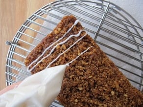 A person holding Greek Yogurt and pouring it on top of the Greek Yogurt Apple Streusel Cake on a cooling rack