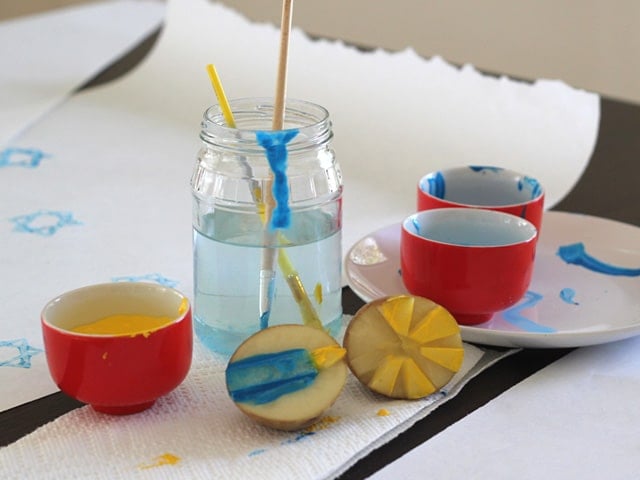 Potato stamps painted with blue and yellow paint, paintbrushes in jar of water, red pots with paint.