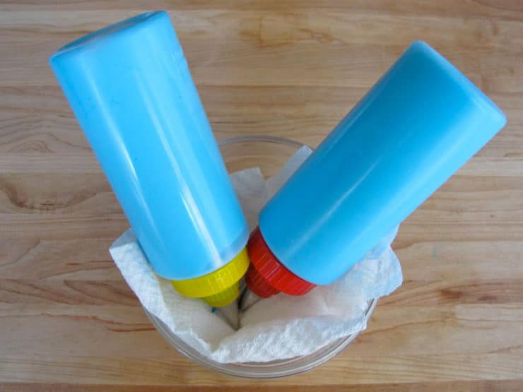 Two icing bottles tip down in bowl lined with wet paper towel.