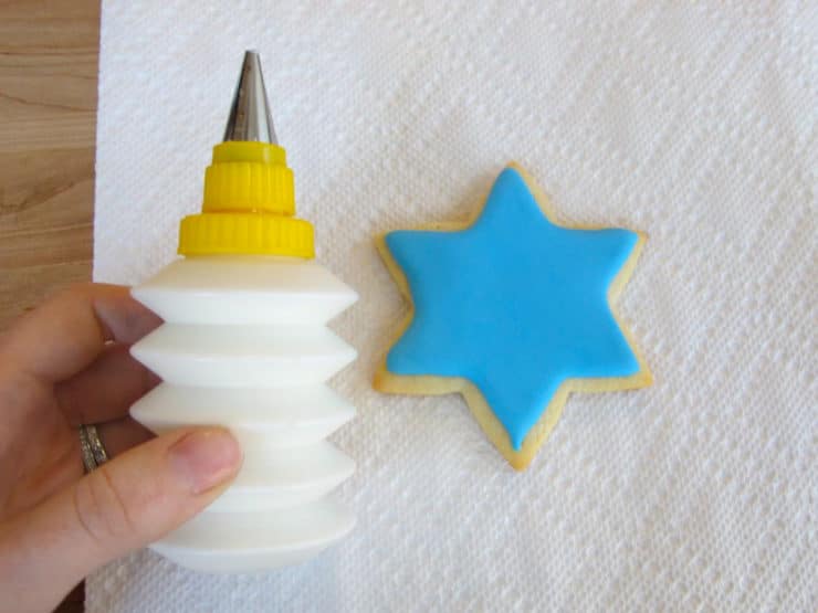 Hand holding small bottle of white royal icing next to blue iced star of David cookie.