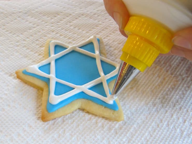 White royal icing linear details being added to blue Jewish holiday sugar cookie.