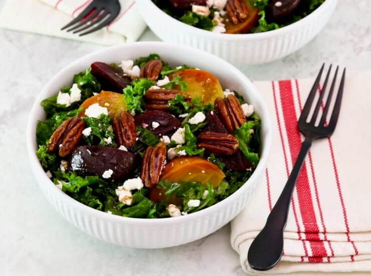 Horizontal shot - shallow bowl filled with roasted beet salad with kale and goat cheese. Fork and striped linen napkin beside the bowl. Partial view of another salad bowl filled with salad, fork, and napkin in background. All resting on a white marble countertop.