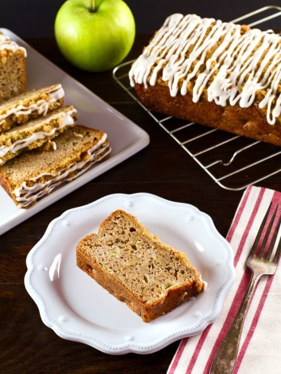 Vertical shot of slice of Greek Yogurt Apple Streusel Cake on white plate with cloth napkin and fork, sliced cake and whole loaf cake on baking rack with green apple in background. 