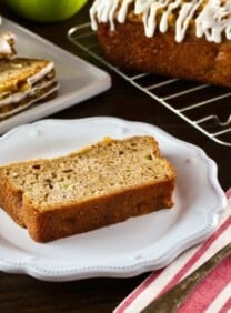 Horizontal shot of slice of Greek Yogurt Apple Streusel Cake on white plate with cloth napkin and fork, sliced cake and whole loaf cake on baking rack with green apple in background.