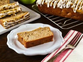 Horizontal shot of slice of Greek Yogurt Apple Streusel Cake on white plate with cloth napkin and fork, sliced cake and whole loaf cake on baking rack with green apple in background.