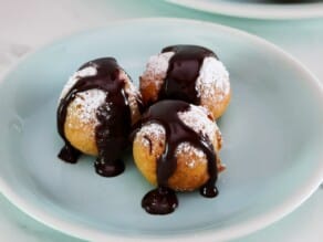 Close up of three buñuelos on a small aqua-colored dessert plate. Buñuelos are dusted with powdered sugar and drizzled with dark chocolate sauce.