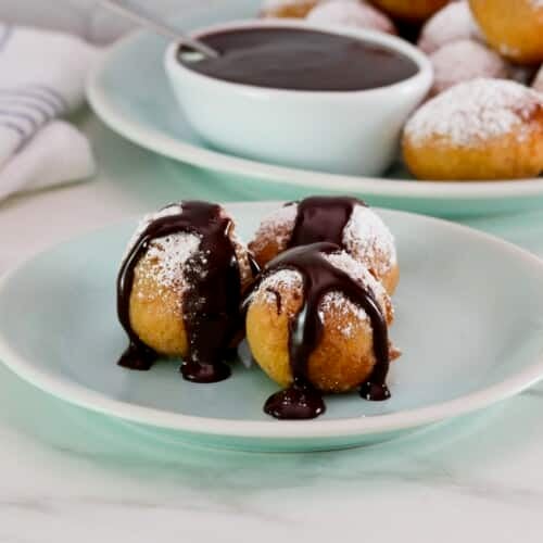 Small dessert plate of three fried buñuelos or bimuelos, topped with powdered sugar and drizzled with chocolate sauce. Platter of powdered sugar buñuelos and a dish of chocolate sauce, cloth napkin in background.