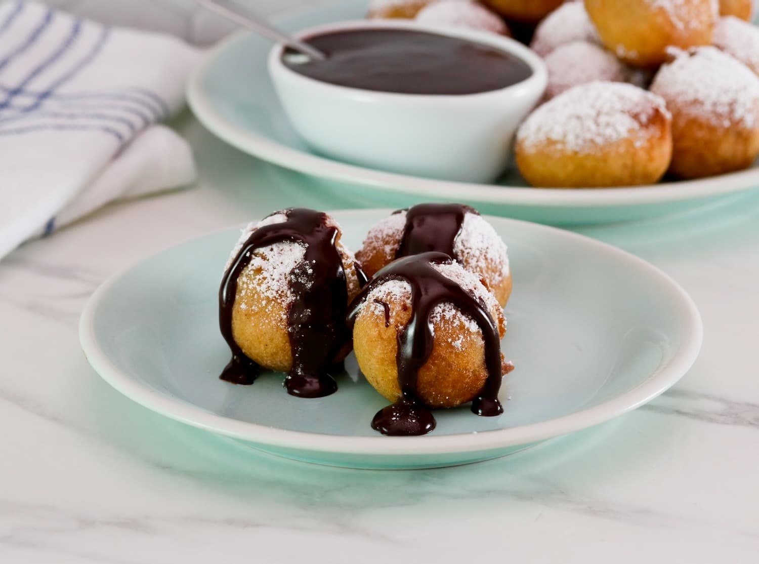 Small dessert plate of three fried buñuelos or bimuelos, topped with powdered sugar and drizzled with chocolate sauce. Platter of powdered sugar buñuelos and a dish of chocolate sauce, cloth napkin in background.