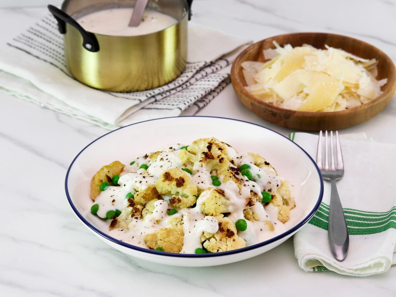 Horizontal shot - large shallow bowl of roasted cauliflower Alfredo with peas, covered in a rich Greek yogurt Alfredo sauce, on a marble countertop. Fork and napkin with green stripes beside the bowl. Small wooden bowl of shaved parmesan in upper right corner of the picture, and a small gold metal pan of Alfredo sauce with freshly cracked black pepper in upper left hand corner. Gold pan rests on a white linen towel.